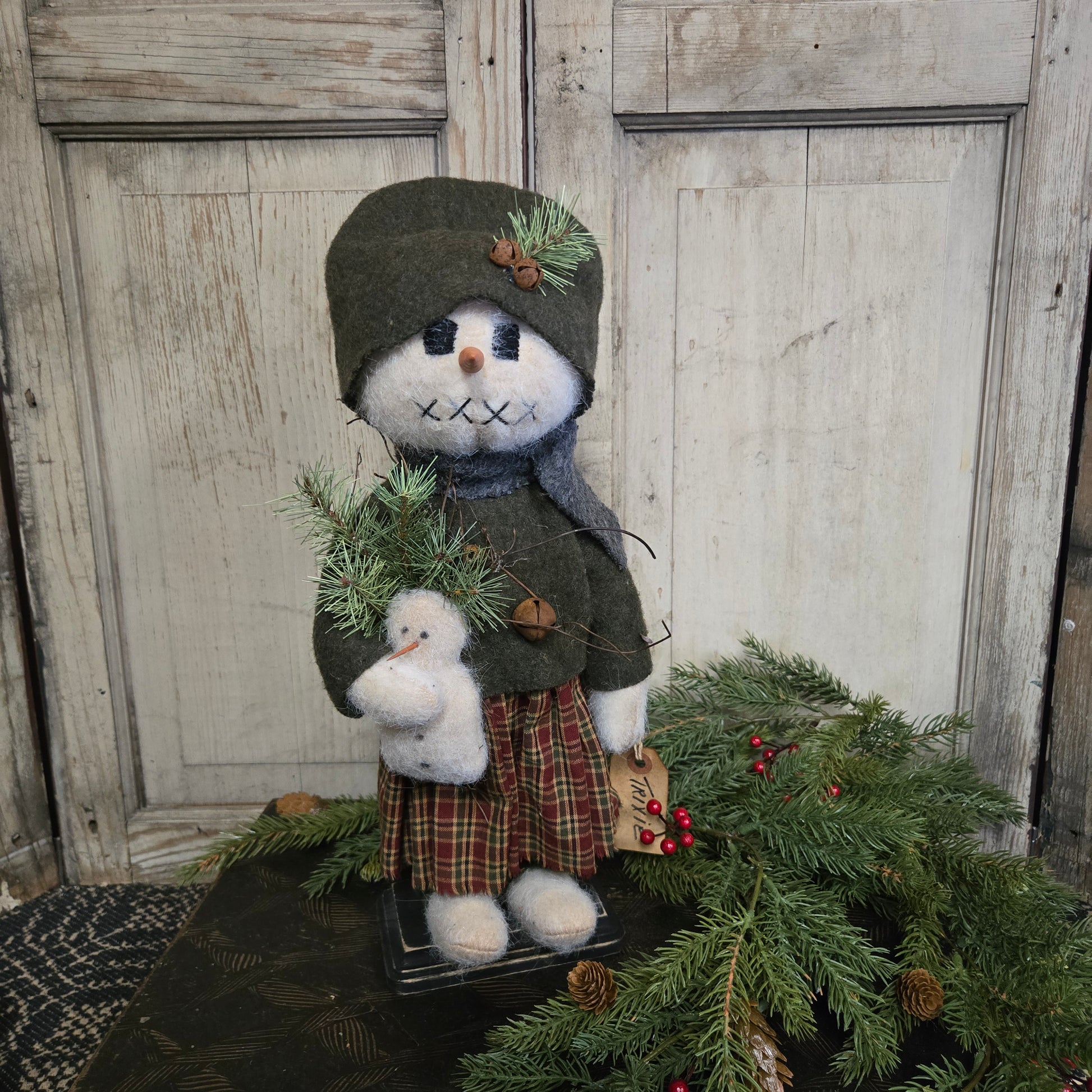 Decorative snowman with a plaid coat and green hat on a wooden surface with a Christmas tree.