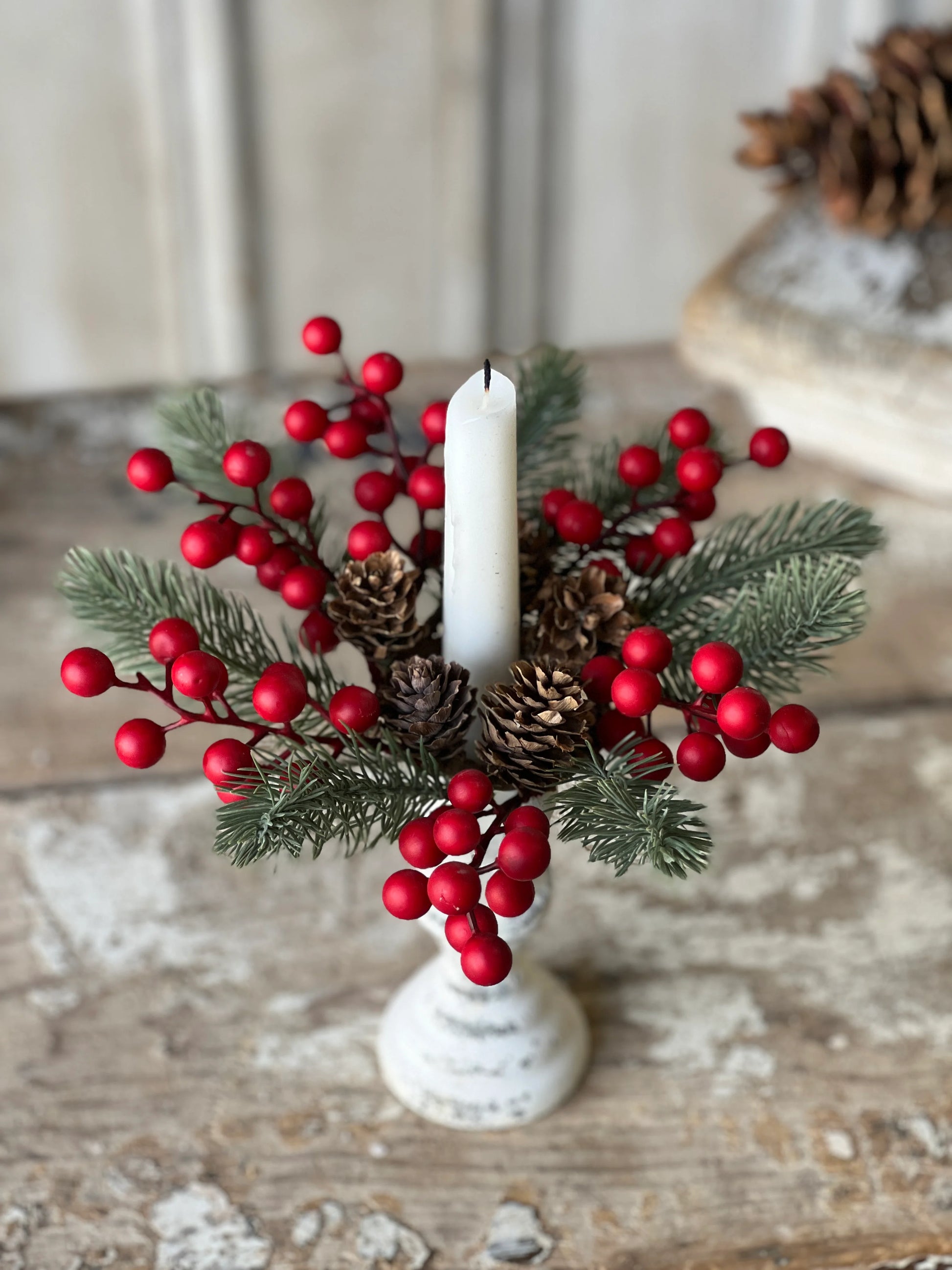 A candle ring with green foliage, red berries, and pine cones, displayed on a white surface.