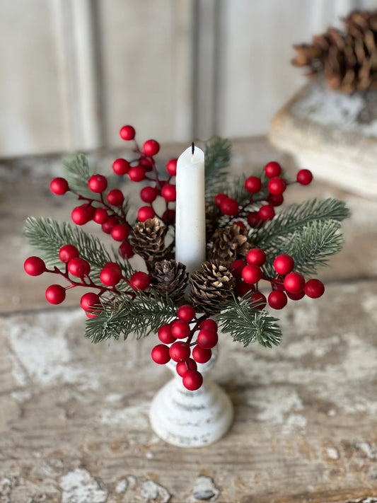 A candle ring with green foliage, red berries, and pine cones, displayed on a white surface.