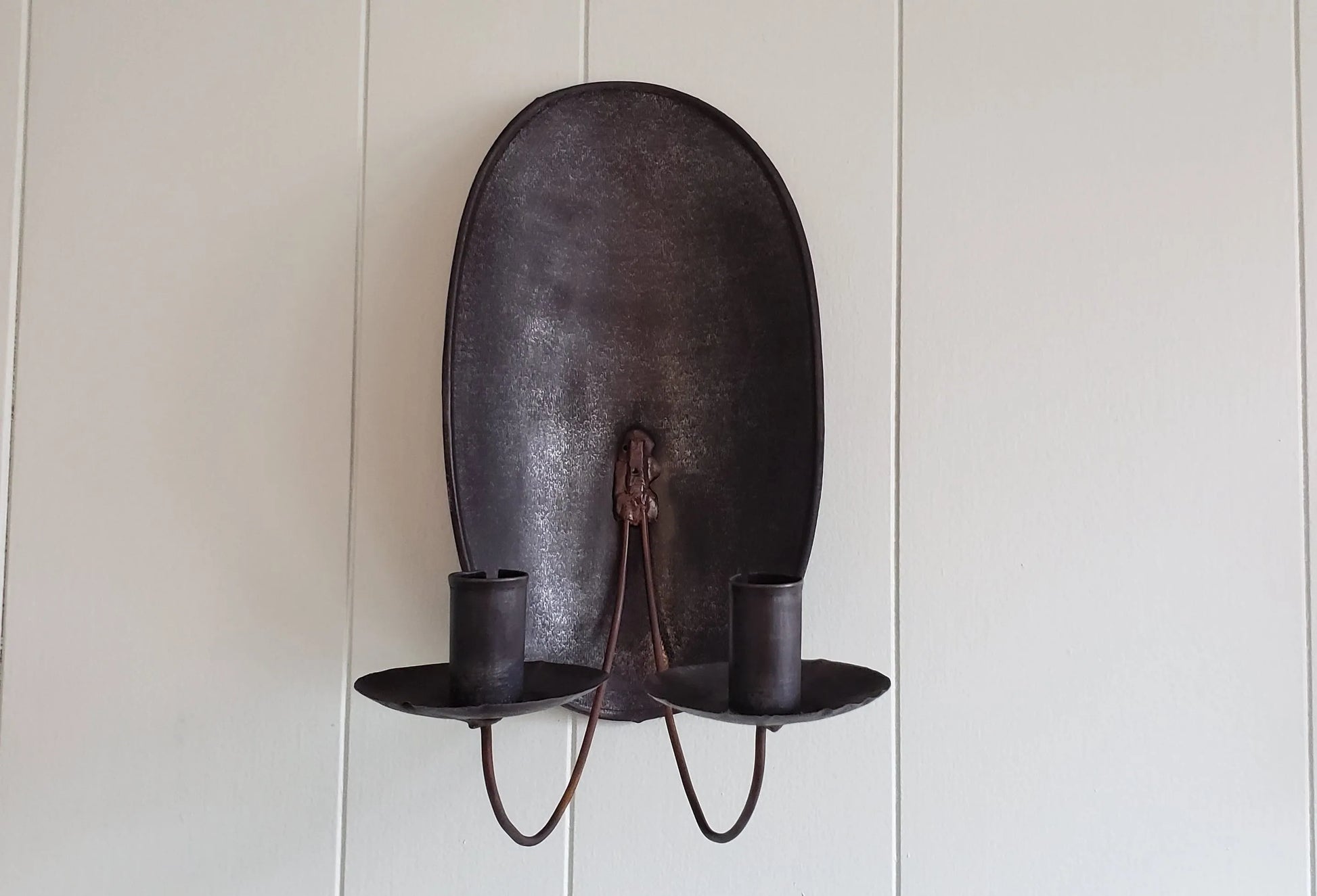 A brown metal turtleback tin candle sconce mounted on a wall, with two candle holders.