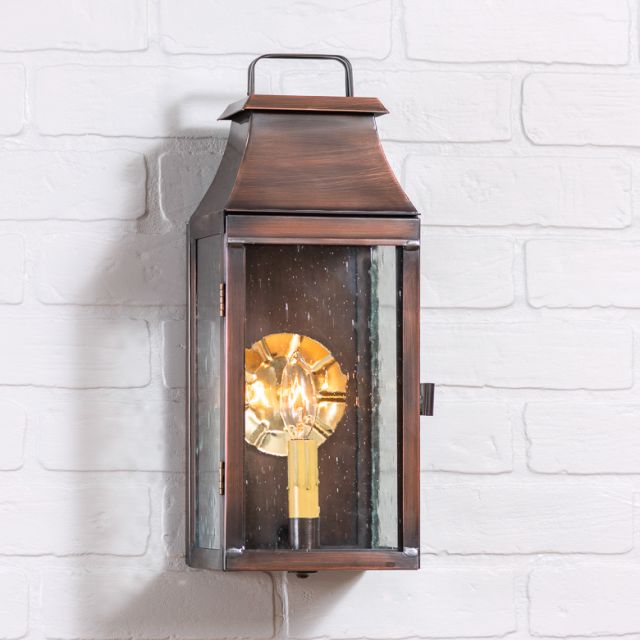 An outdoor wall-mounted lantern with a solid antique copper finish, blackened solder, and a clear glass front panel, displaying a single light bulb.