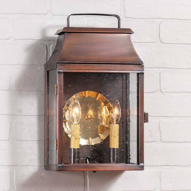 An outdoor wall light fixture with a solid antique copper finish, featuring a blackened solder and lacquer coating, mounted on a white brick wall. It has a reflector with two bulb holders inside and is hung by a metal bracket.