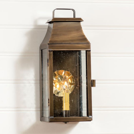 Outdoor wall-mounted lantern with a solid weathered brass finish, featuring a clear glass front and a single light bulb inside, mounted on a white wall.