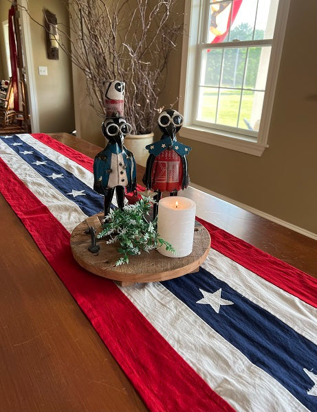 A vintage-style table runner with a flag pattern displayed on a dining table, flanked by two decorative figurines and a centerpiece.