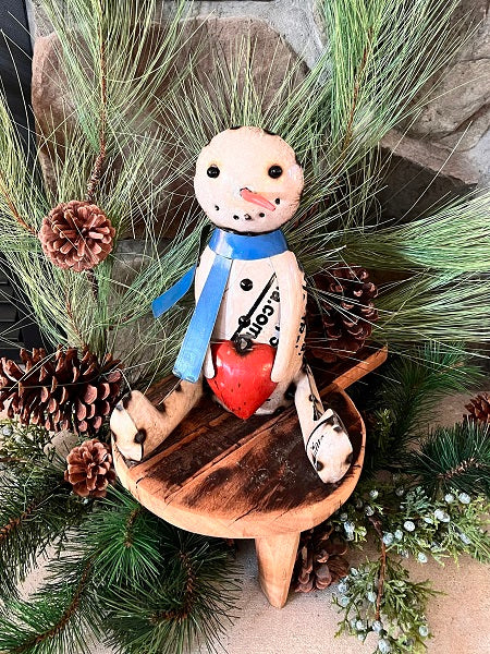 A handcrafted metal snowman figurine with a blue scarf and a red heart, sitting on a wooden stool, surrounded by green foliage and pine cones.