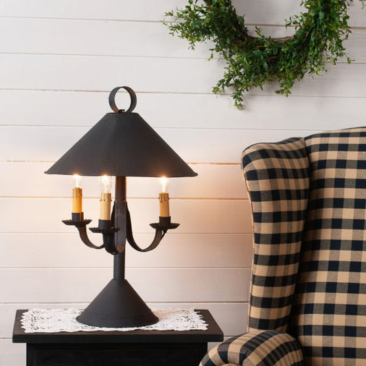 A textured black metal lamp with a classic shade, featuring three sockets for adjustable brightness, placed on a table next to a plaid armchair.