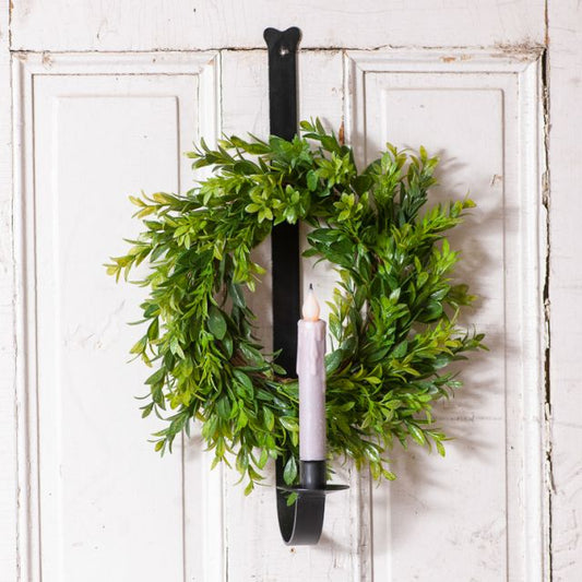 A wall mount wreath and candle holder with a green wreath hanging on a white door and a black candle holder attached to the wreath.