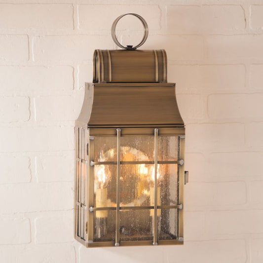 A weathered brass wall lantern with seedy antique glass and a visible light source, mounted on a white brick wall.