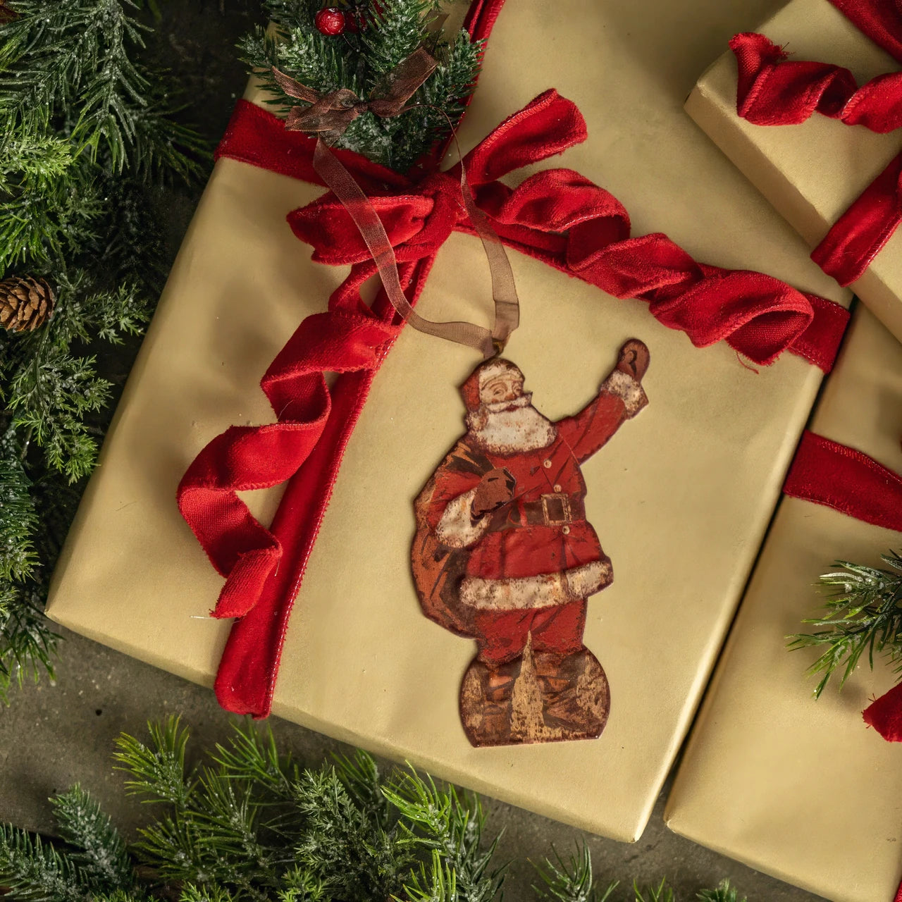 A vintage-style Santa Claus hanging ornament attached to a red ribbon bow on a gold gift box, surrounded by pine branches and other wrapped presents.
