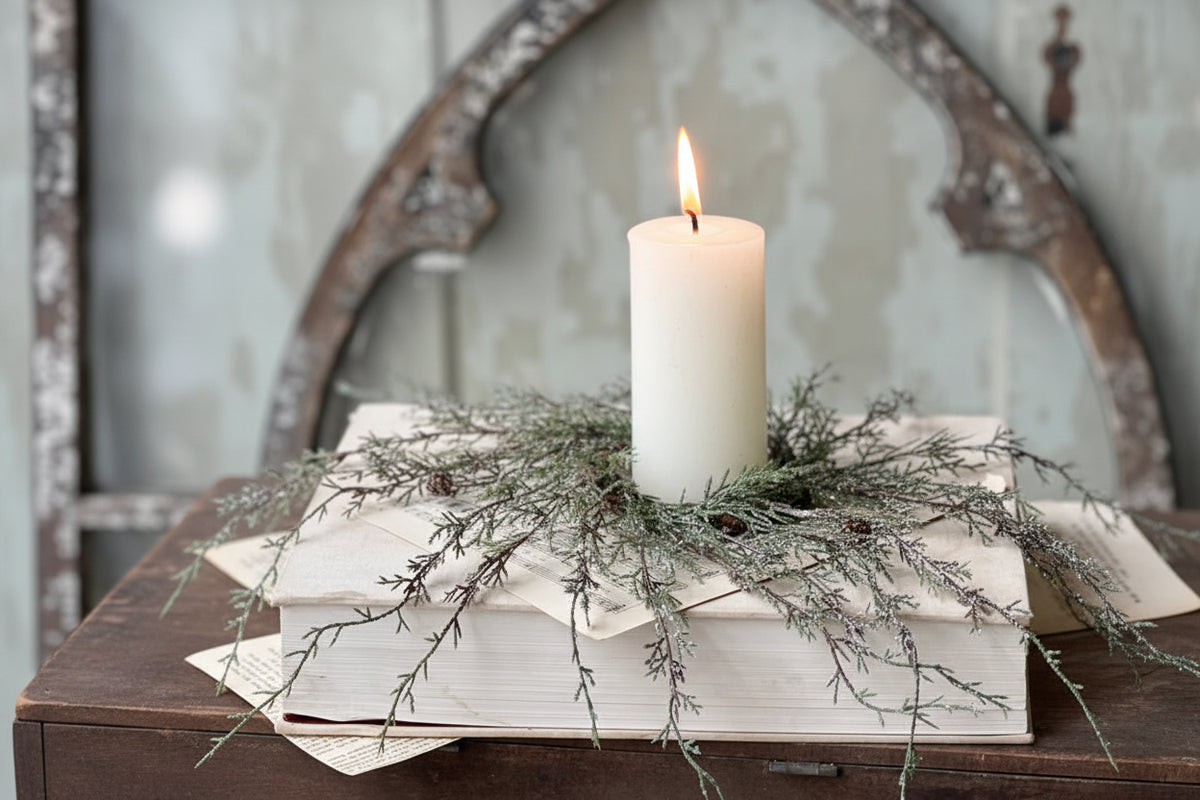 White candle on a wooden surface with greenery and books, rustic decor.