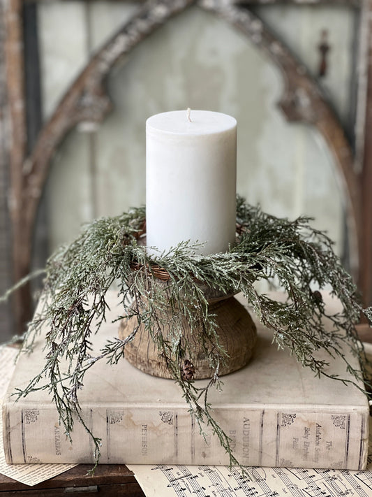 A candle ring featuring greenery and pine cones, dusted with snow and glitter, displayed around a white candle.
