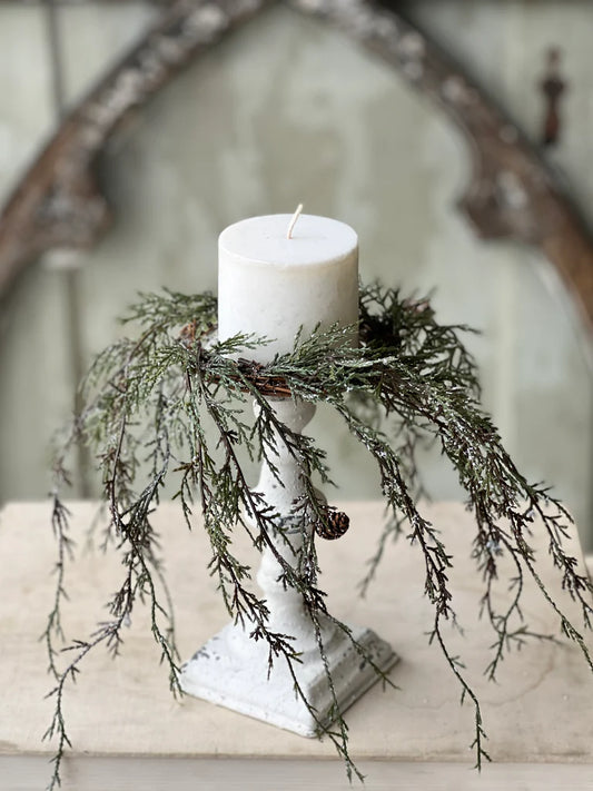 Weeping cedar candle ring with snow and pinecones on a white pedestal holding a white pillar candle.