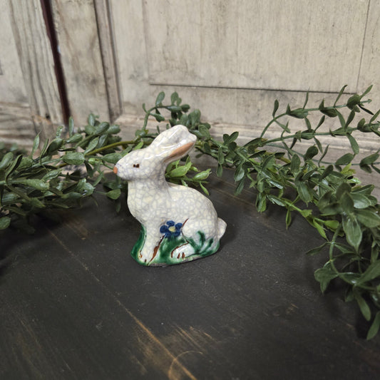 Pottery sculpture of a white rabbit with a blue flower standing on a black wood surface surrounded by greenery