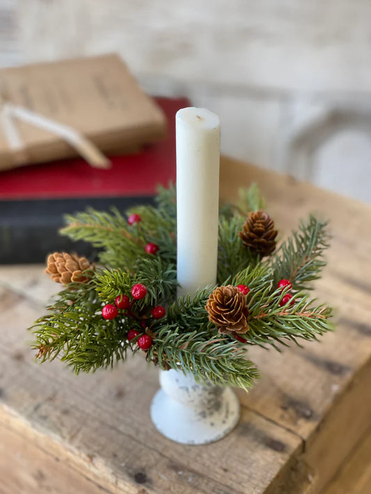 A candle ring with white spruce branches, red berries, and pine cones, displayed around a white candle, placed on a wooden surface.