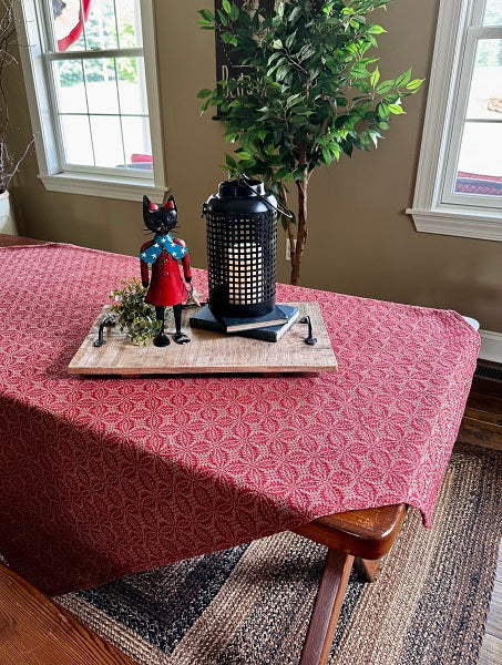 A red patterned tablecloth draped over a wooden dining table in a home setting, styled with a decorative tray, a black lantern, and a small figurine. The background features a beige wall, windows, and a potted green tree.