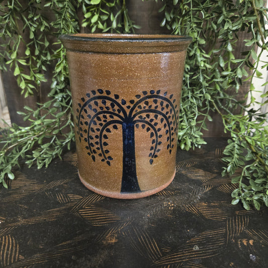 Stoneware crock with decorative willow tree design on a textured surface with greenery