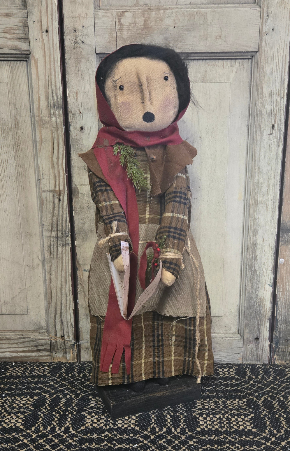 A festive holiday figurine of a female character in a red and brown checkered dress, holding a song book, with a wooden backdrop.