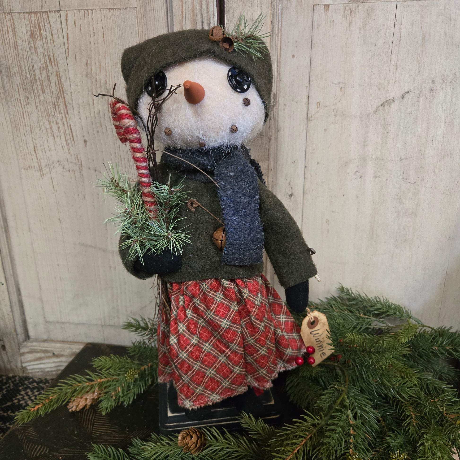 Decorative snowman figurine with a plaid coat and green hat on a wooden surface with Christmas tree branches.