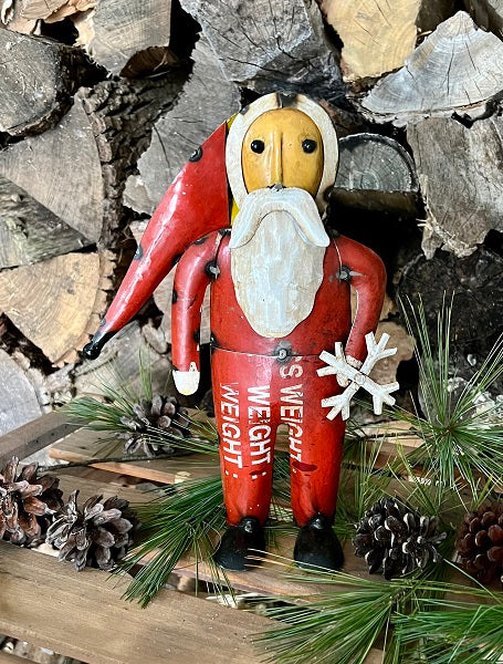 A wooden Santa figurine with a red outfit and white beard, holding a pair of snowshoes, positioned against a backdrop of pine logs and decorative pine cones.