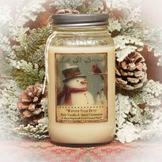 A mason jar candle with a label featuring a vintage-style snowman, pine cones, and the scent marked as 'Winter Star Dust'. The candle is placed in front of a backdrop of pine branches and decorative fabric.