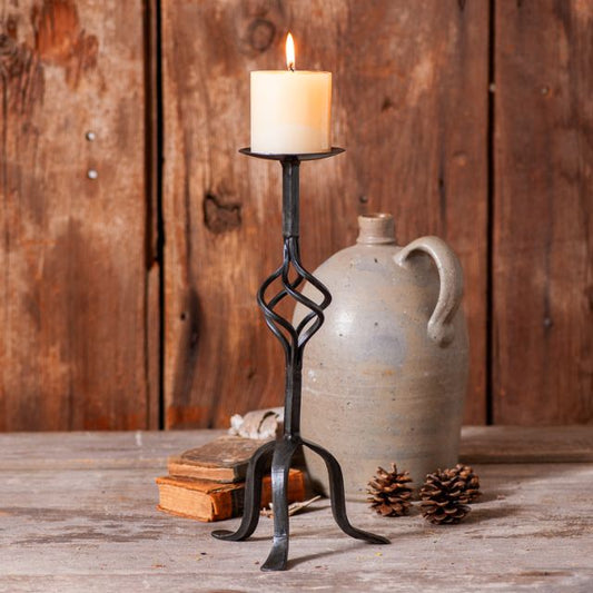 A wrought iron pillar candle holder with an elegant twisted basket on the center post, placed on a wooden surface next to a ceramic jug and pine cones.