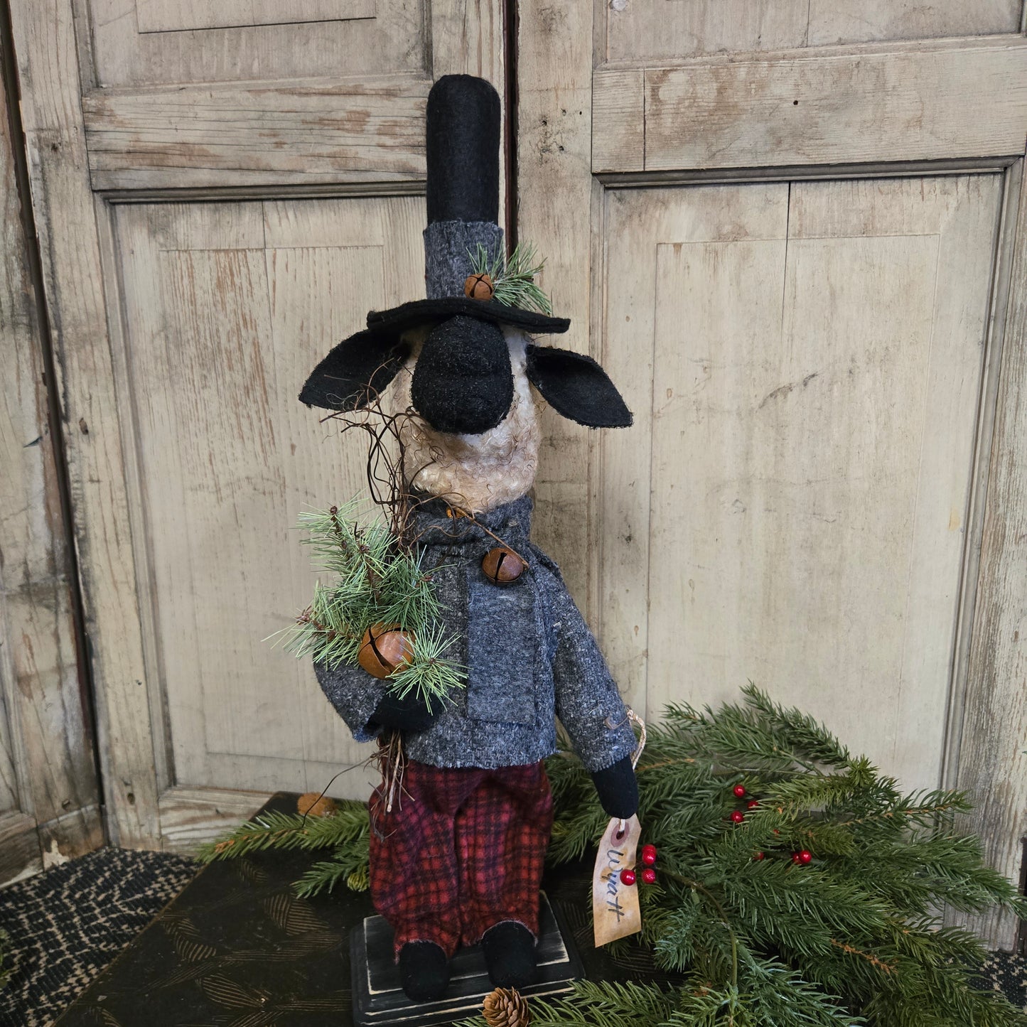 Decorative figure of a sheep wearing a coat and hat, holding a wreath against a wooden door.