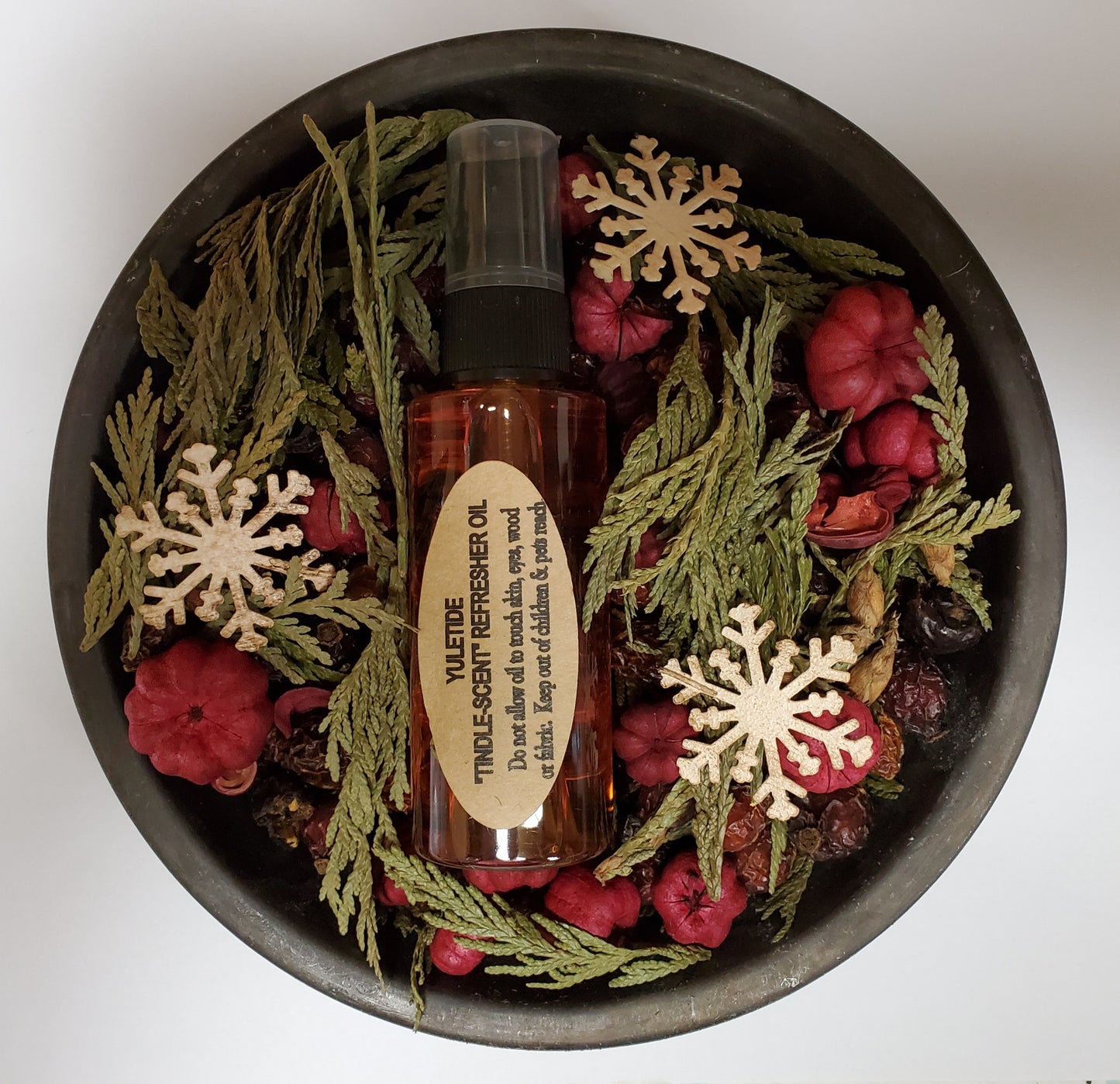 A metal pan with a glass bottle labeled 'Yuletide Tindle' surrounded by decorative potpourri, pine cones, and red and brown berries.