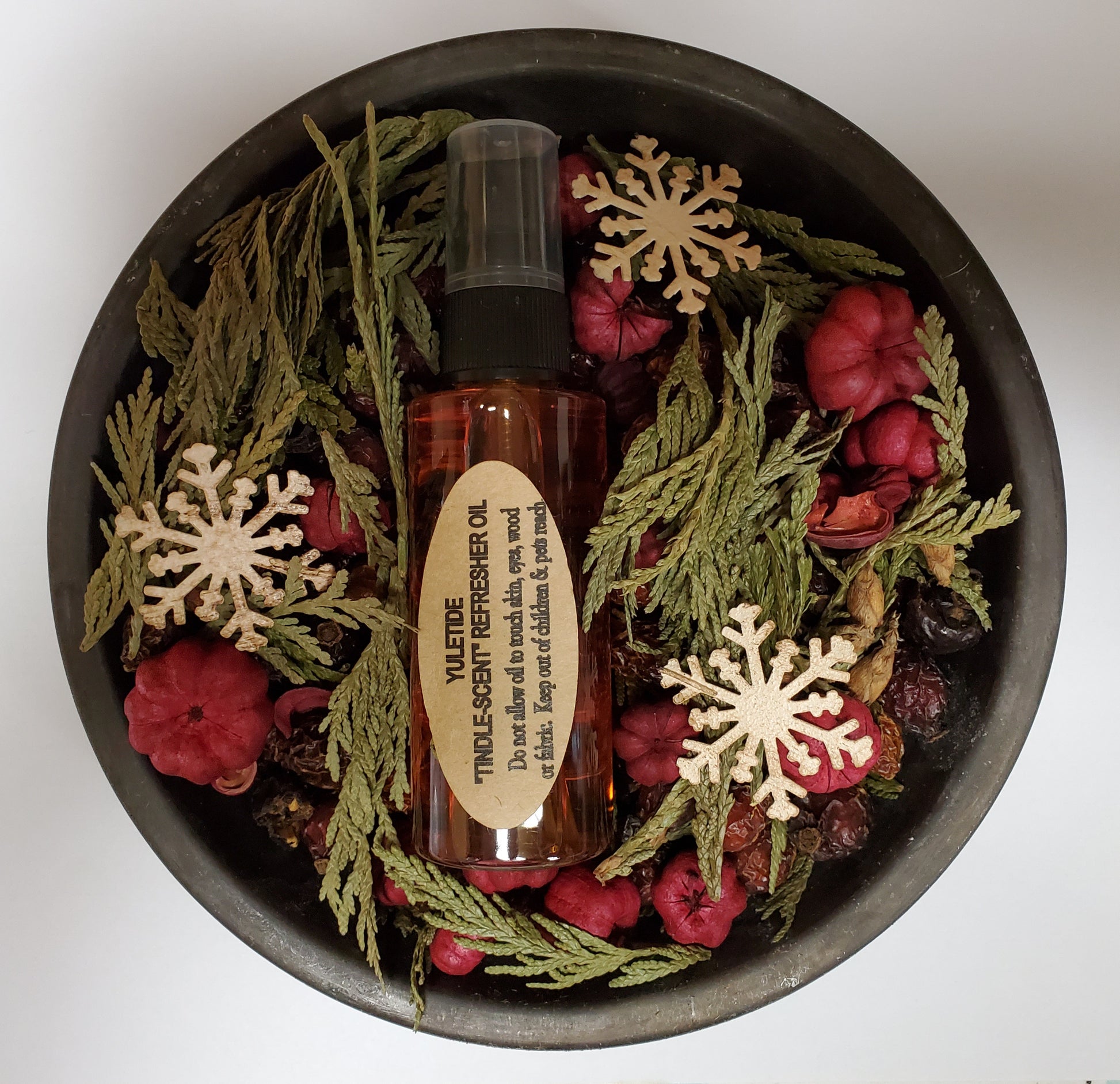 A metal pan with a glass bottle labeled 'Yuletide Tindle' surrounded by decorative potpourri, pine cones, and red and brown berries.