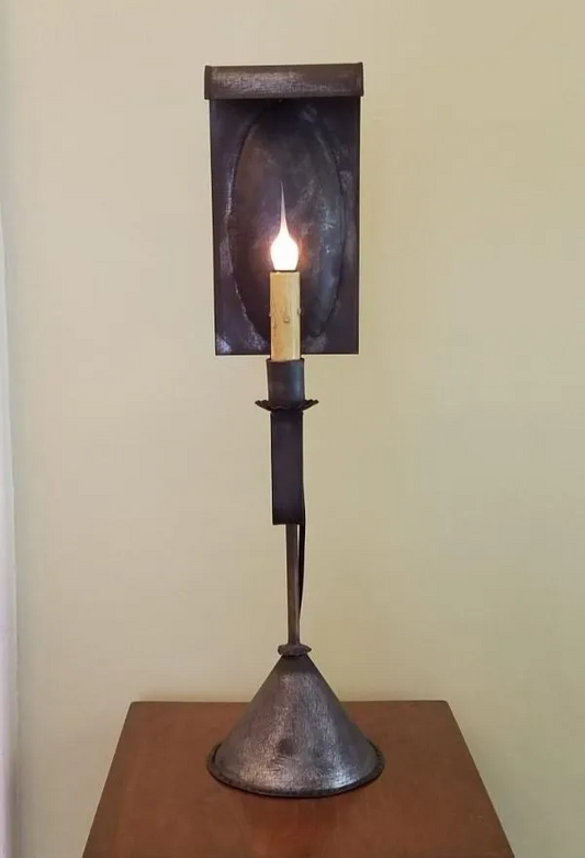 A Zoar Tin Table Light with a lit candle, featuring a metallic bronze-colored base and a black candle holder, placed on a wooden surface against a beige wall.